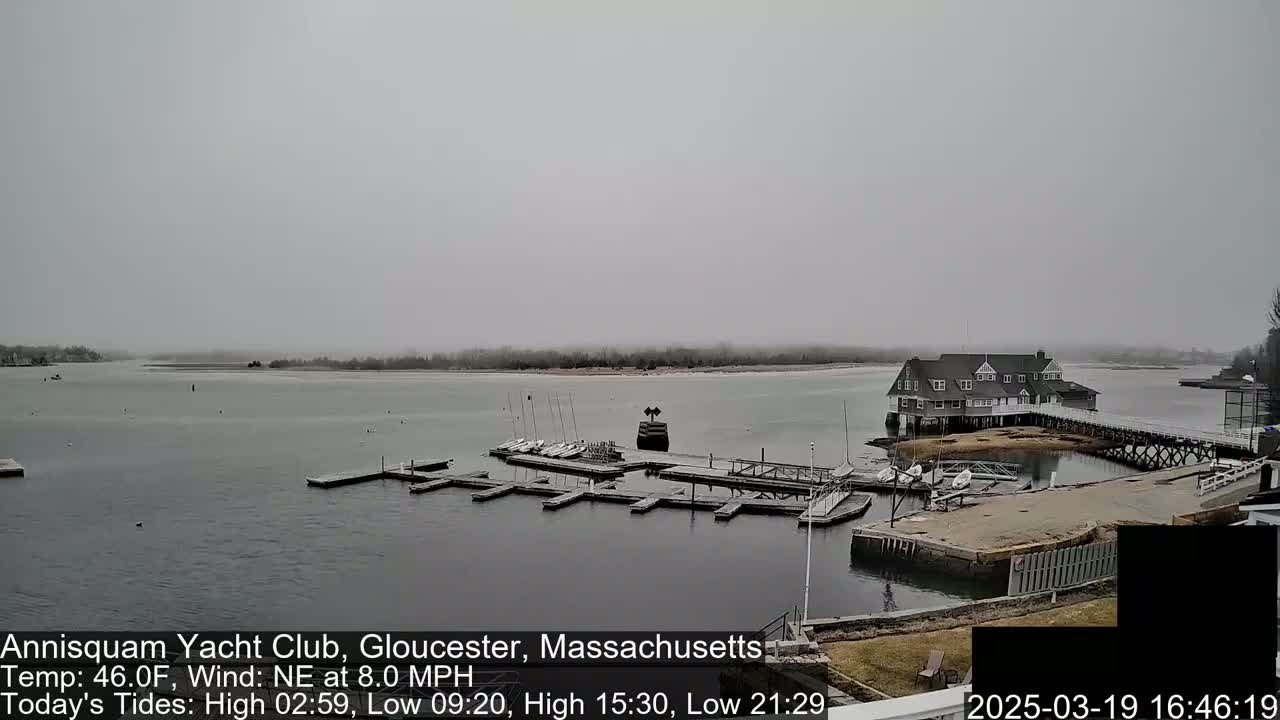 A hazy, overcast day shows a marina with sailboats docked at a clubhouse on a calm body of water.
