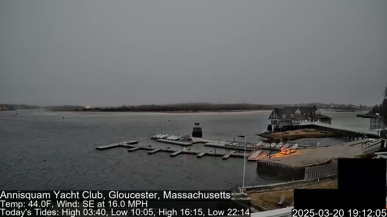 A mostly cloudy, overcast day shows a marina with many small sailboats docked, a large clubhouse on an island, and calm water under a gray sky.