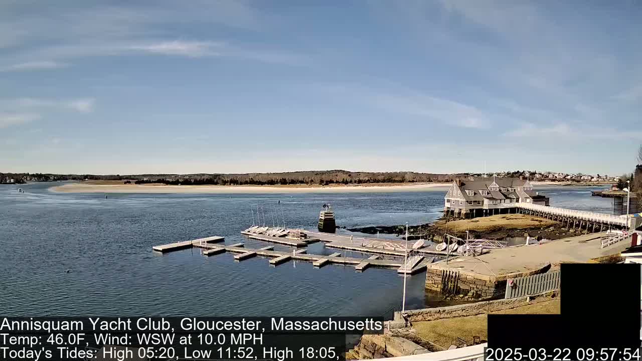 A marina with numerous boats docked at a clubhouse, situated on a calm body of water under a mostly sunny sky.