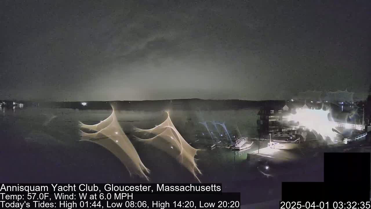 A nighttime, waterfront scene shows a body of water under a cloudy sky with bright lights illuminating a building and dock area, and streaks of light possibly caused by long exposure photography or lightning.