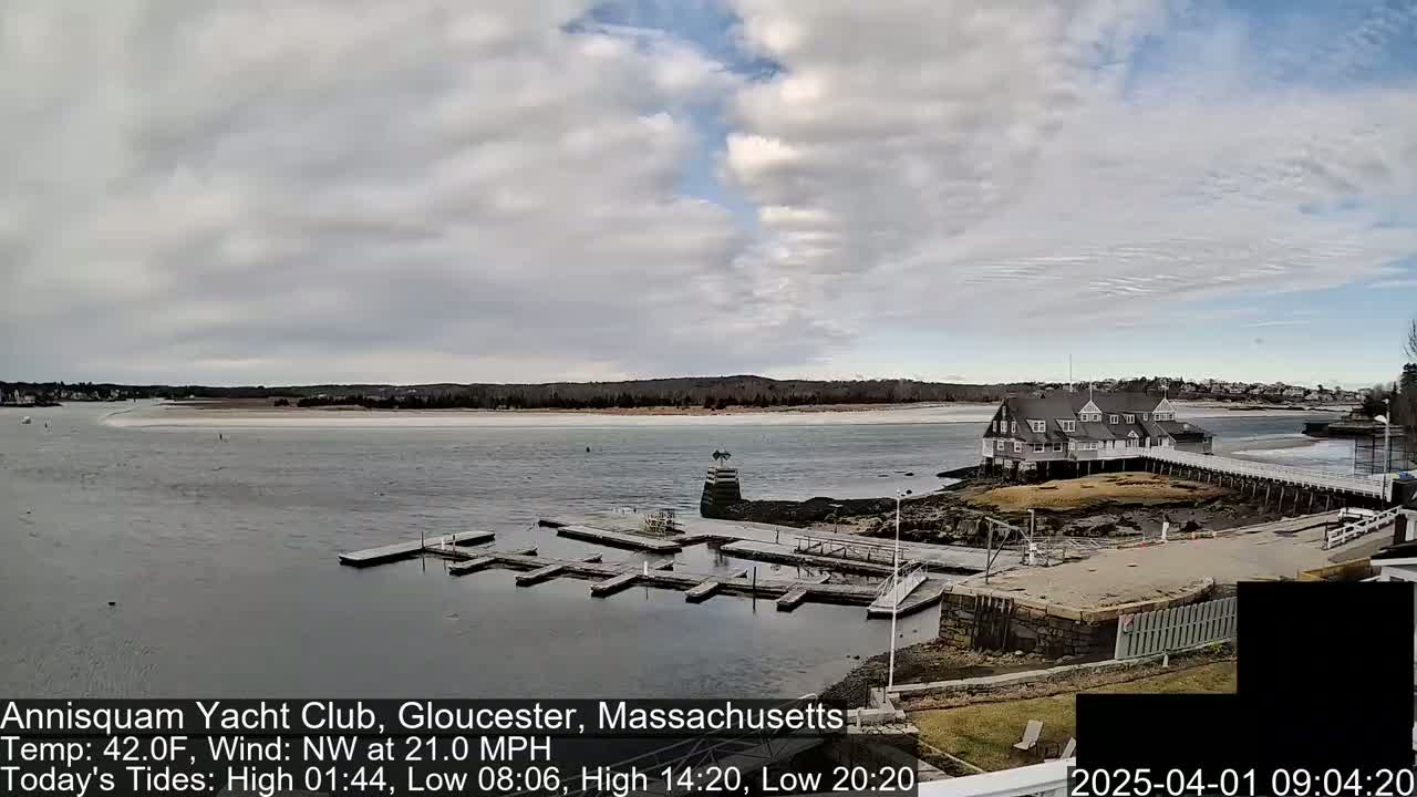 A cloudy day shows a waterfront view of a yacht club with a dock, surrounded by calm water and a sandy shore under a partly cloudy sky.