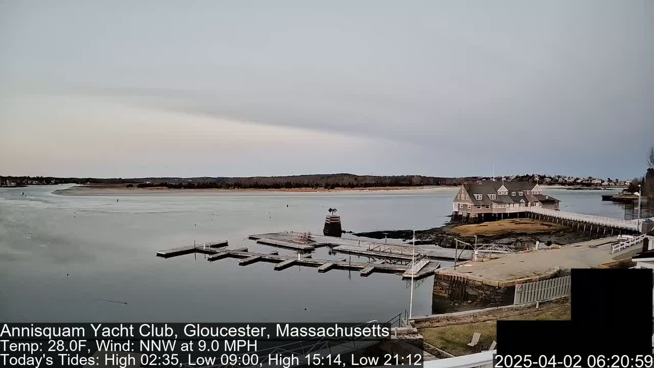 A calm bay with a small clubhouse on a pier, empty boat slips, and a partially frozen shoreline under an overcast sky.