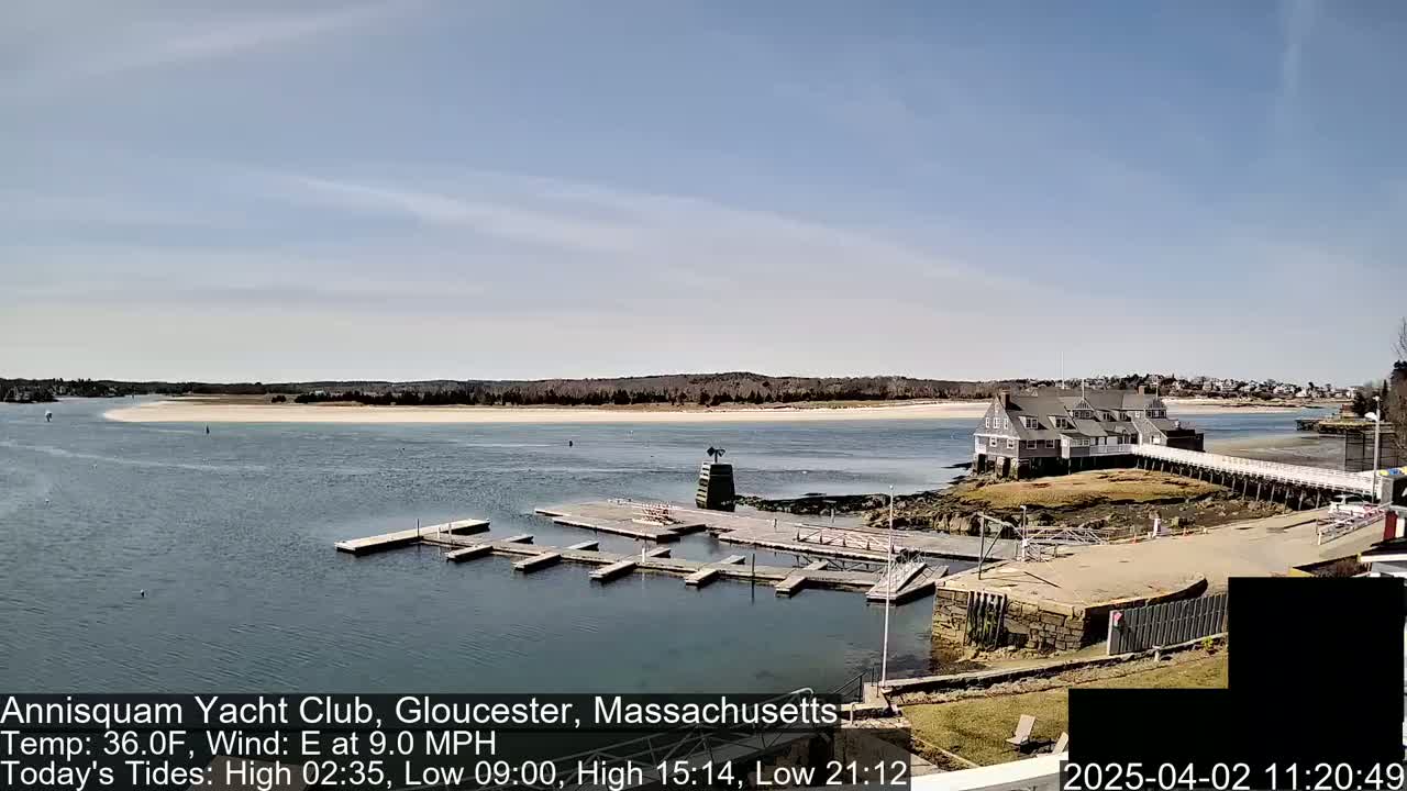 A yacht club sits on a small island connected to the mainland by a pier, with a sandy beach visible across a calm body of water under a mostly clear sky.