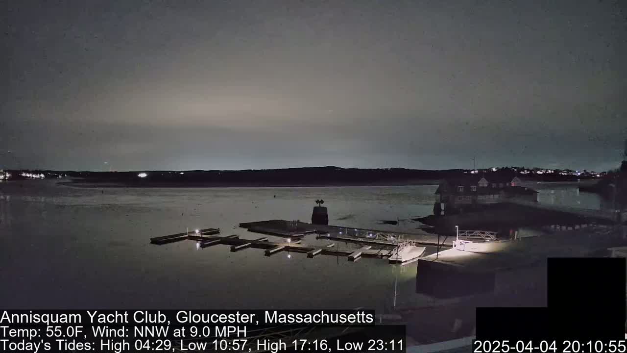 A nighttime view of a calm body of water with a marina and a building on the shore under a dark, overcast sky.