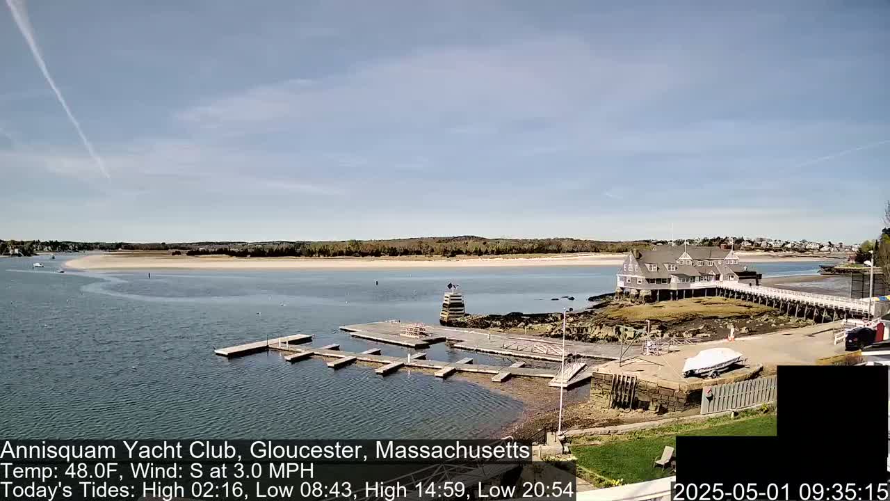 A yacht club sits on a rocky outcropping next to a sandy beach, with calm water and a partly sunny sky.