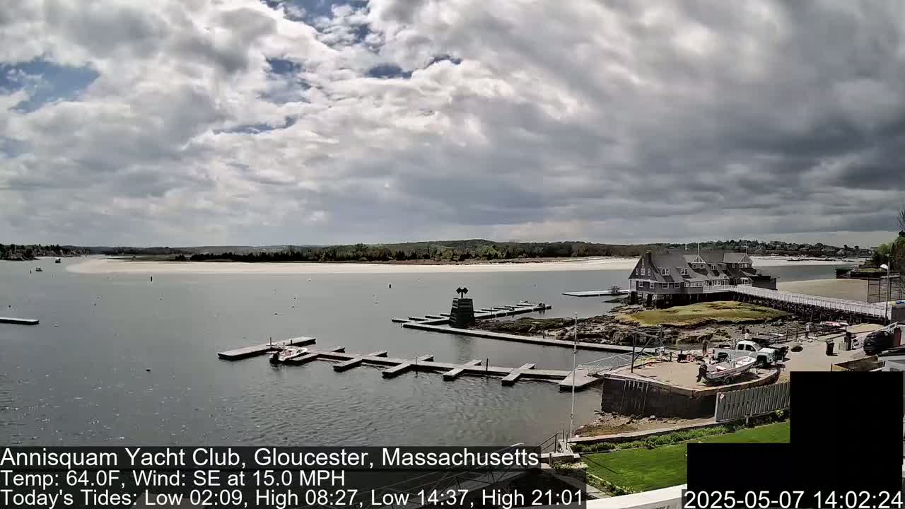 A mostly cloudy day shows a marina with boats and a clubhouse situated on a rocky peninsula next to a sandy beach.
