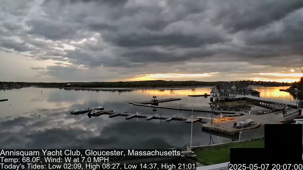 A calm body of water with docked boats and a large building is under a partly cloudy sky with a sunset.