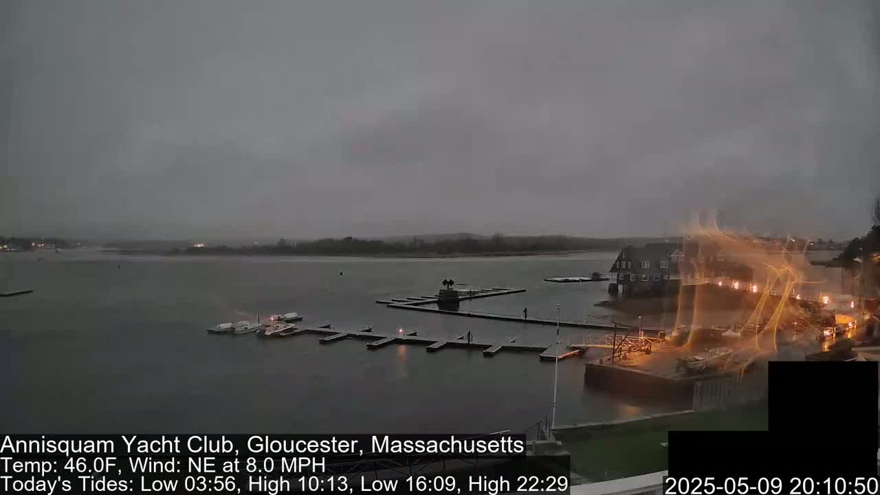 A mostly overcast, gray day shows a marina with boats moored at docks, a clubhouse, and calm water under a northeastern wind.