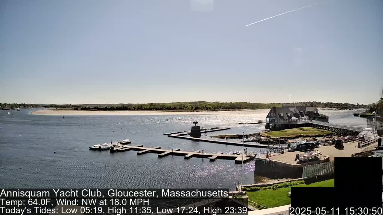 A calm, sunny day shows a marina with boats docked, a clubhouse on a small island, and a sandy beach across a waterway under a mostly clear sky.