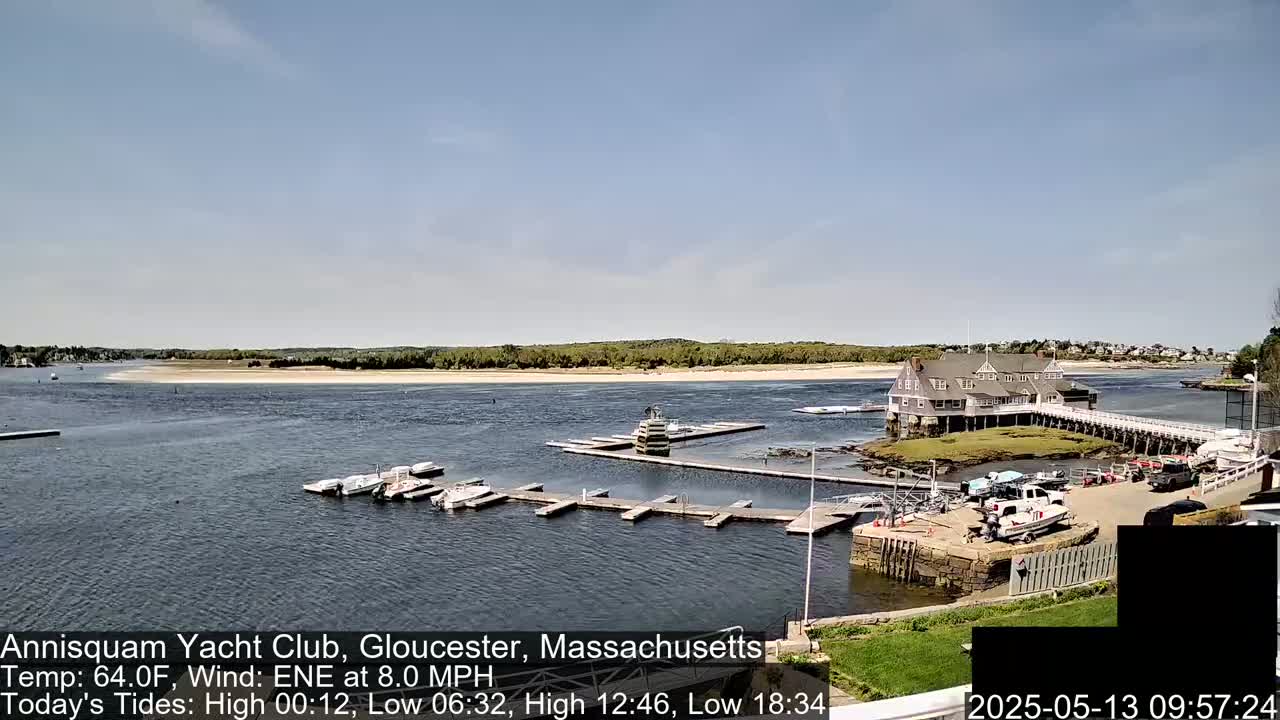 A calm body of water shows a sandy beach, a large building on a small island, several boats docked at piers, and a clear blue sky.