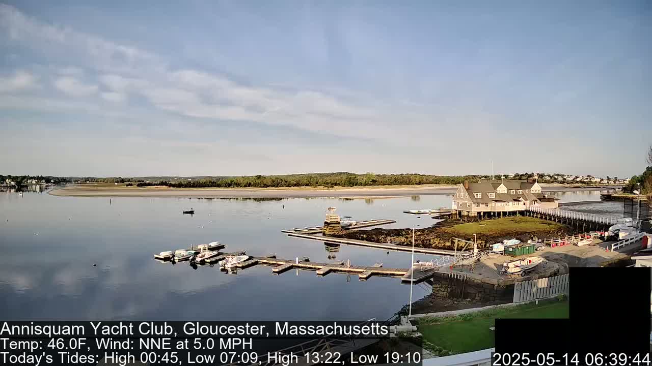 A calm, partly cloudy day shows a yacht club on a small island in a bay, with numerous boats docked at the piers.