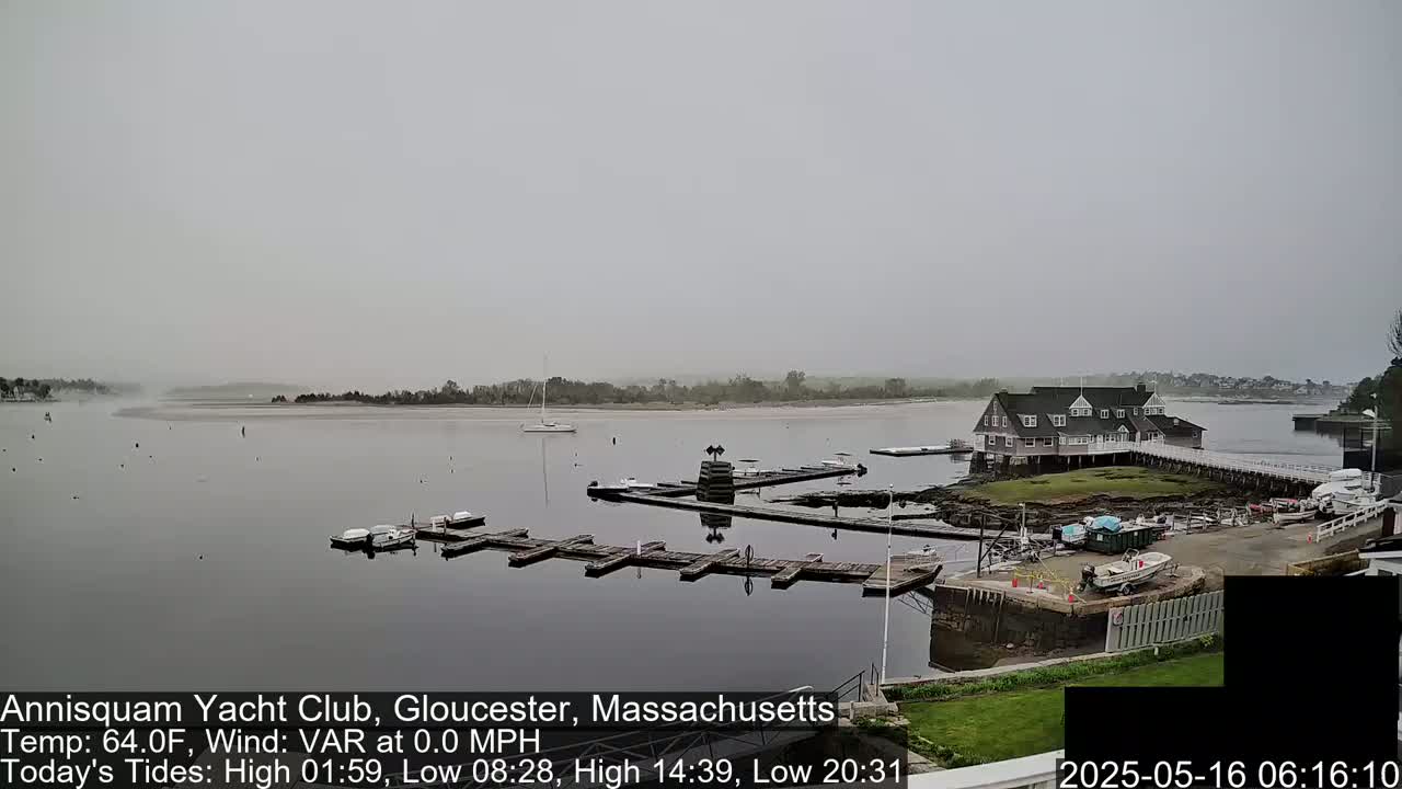 A hazy, calm day reveals a harbor with boats at docks near a clubhouse, and a sandy shore across a calm body of water.