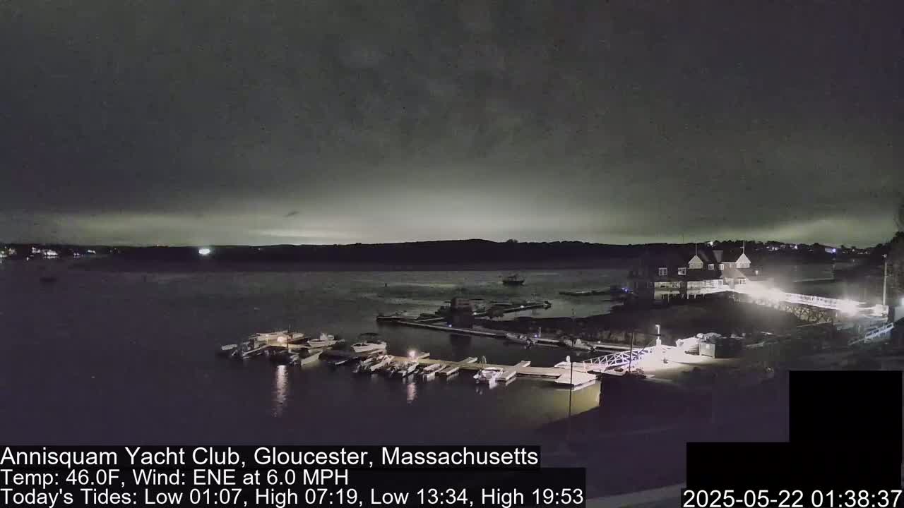 A nighttime view of a marina with boats docked, a clubhouse, and a bridge, under a dark, cloudy sky.