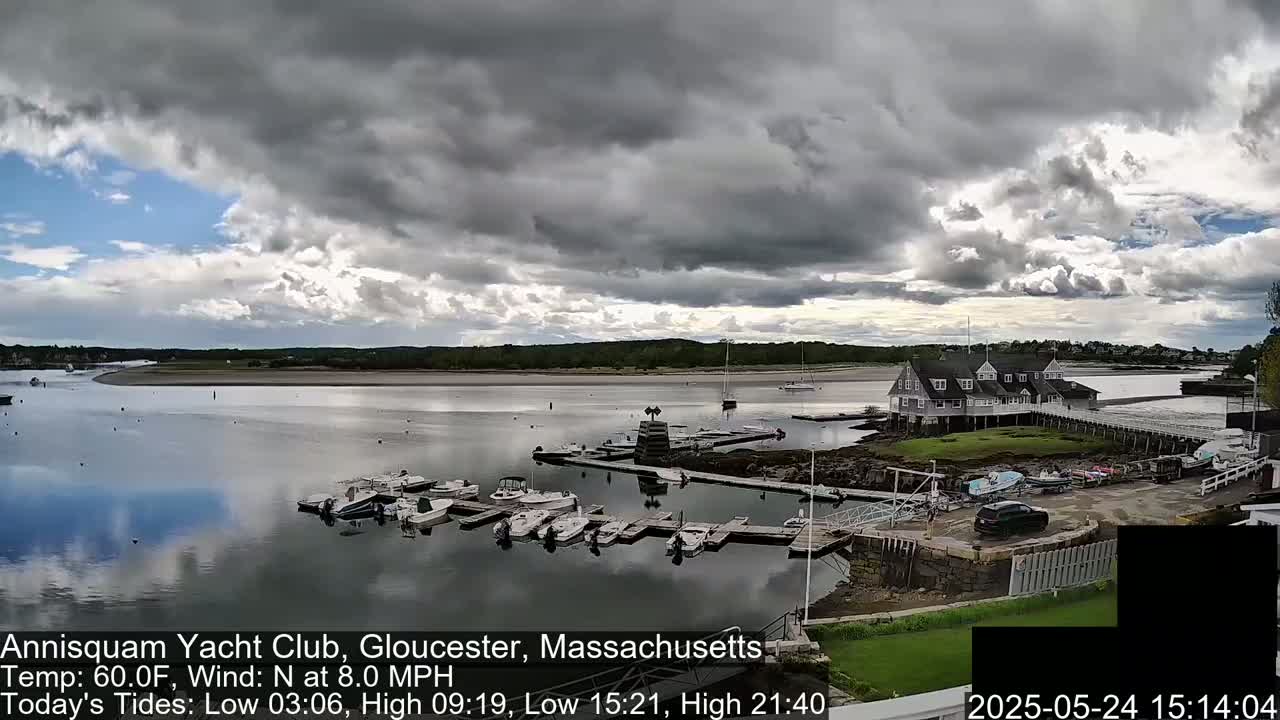 A marina filled with boats sits under a partly cloudy sky, with a calm body of water reflecting the clouds and boats.