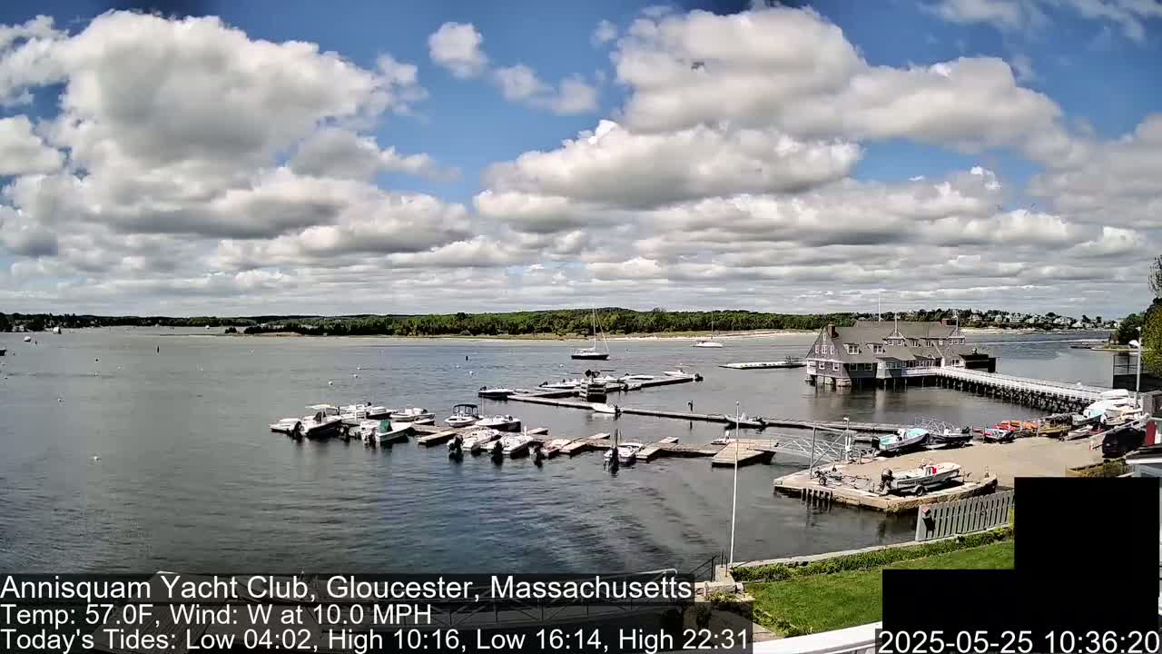 A partly cloudy day shows numerous boats moored at a marina next to a large clubhouse, with additional boats visible on the water.
