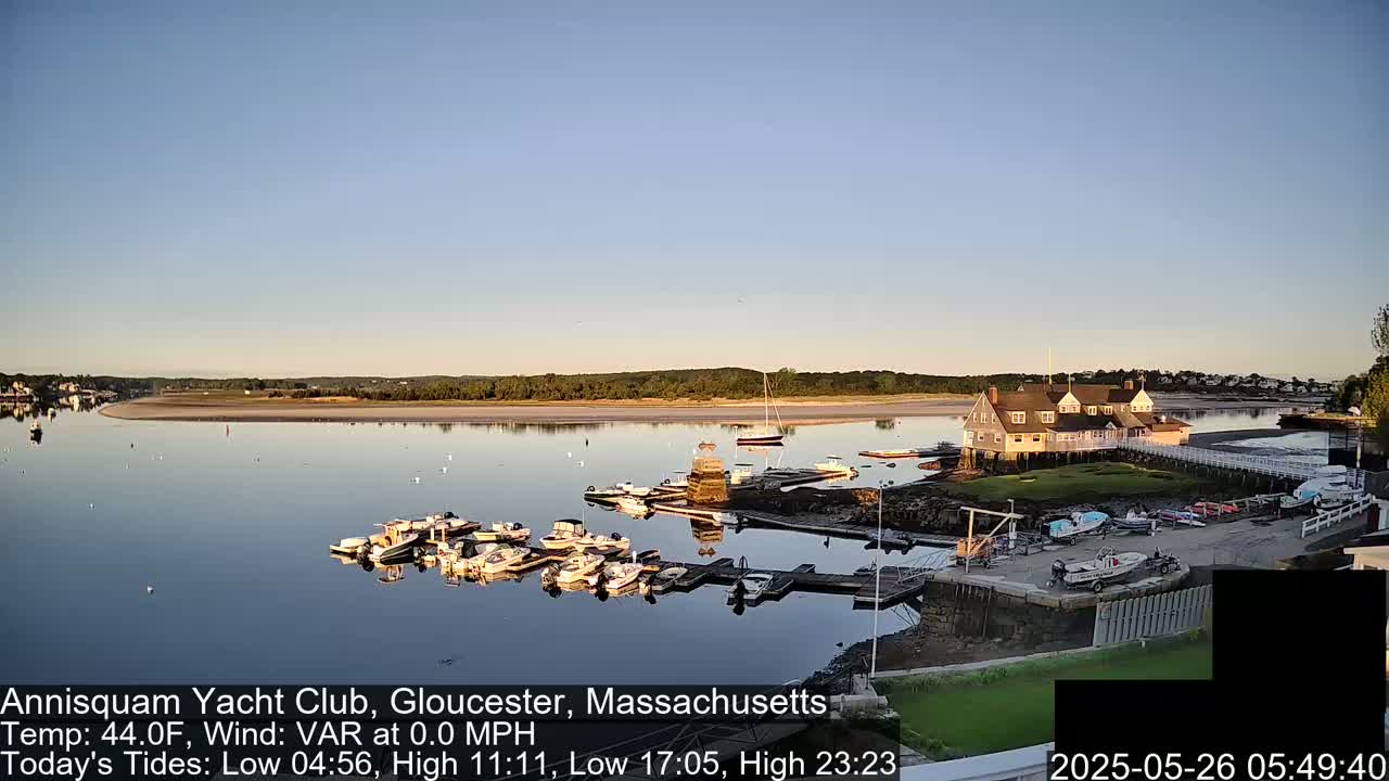 A calm, clear morning reveals a marina filled with boats near a clubhouse situated on a tranquil body of water next to a sandy shore.