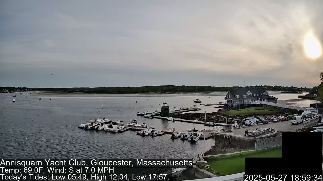 A calm body of water with numerous boats docked at a marina, a clubhouse situated on a small peninsula, and overcast skies.
