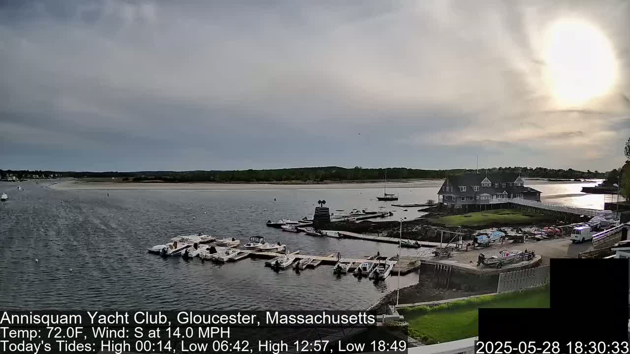 A yacht club sits on a small island in a calm bay, with numerous boats docked nearby under a partly cloudy sky.