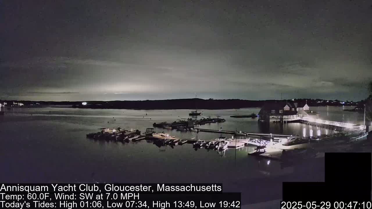 A nighttime view of a marina with boats docked, a clubhouse, and a calm body of water under a mostly cloudy sky.