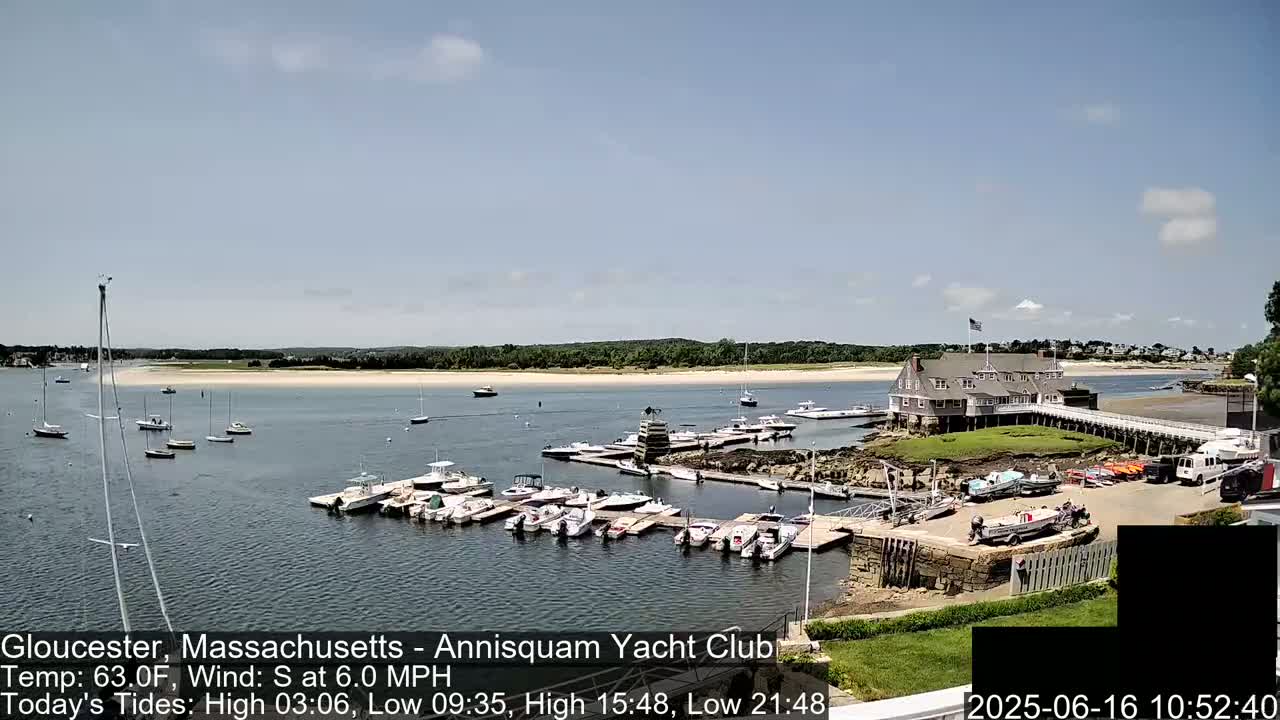 A sunny day shows a harbor filled with numerous boats docked at a yacht club, with a sandy beach and a building in the background.