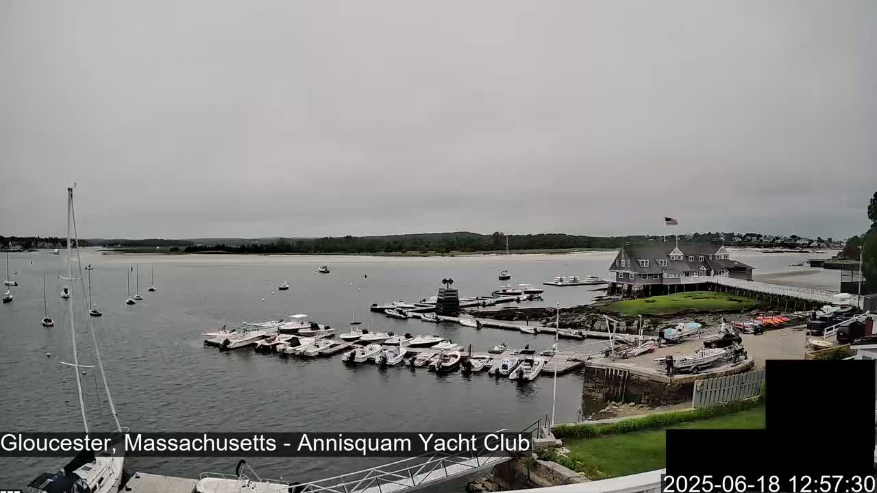 A harbor filled with numerous boats and yachts is seen under an overcast sky, with a waterfront clubhouse visible in the background.
