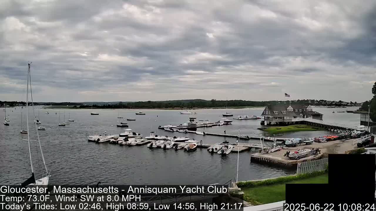 Numerous boats are docked at a marina under a cloudy sky, with a clubhouse visible in the background.
