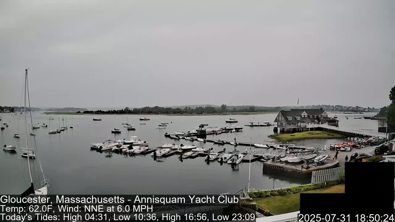 A calm, overcast day shows numerous boats docked at a yacht club on a body of water, with additional boats anchored in the bay.