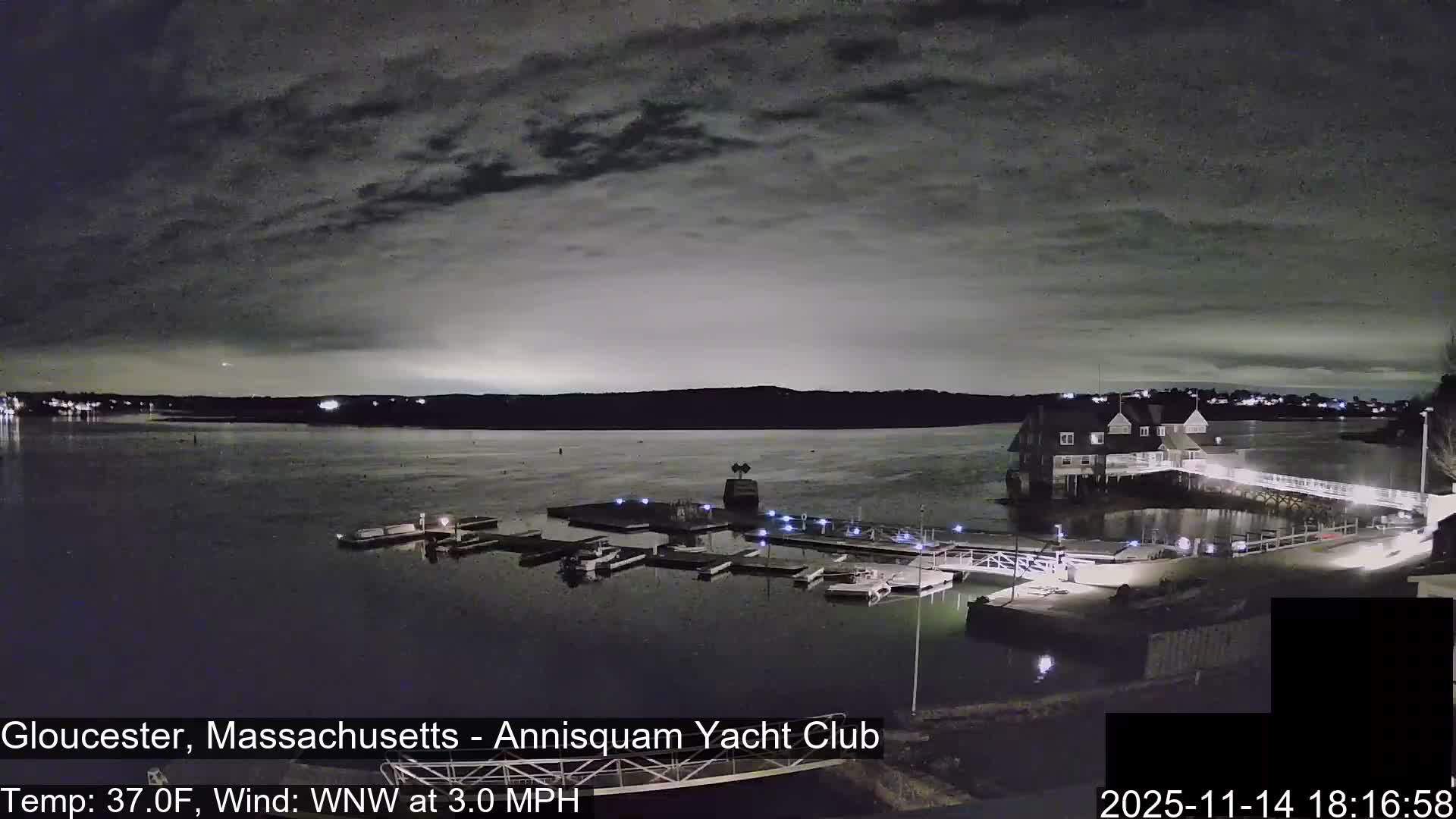 A nighttime scene reveals a calm body of water reflecting lights from docks, boats, and a yacht club building under a mostly cloudy sky.