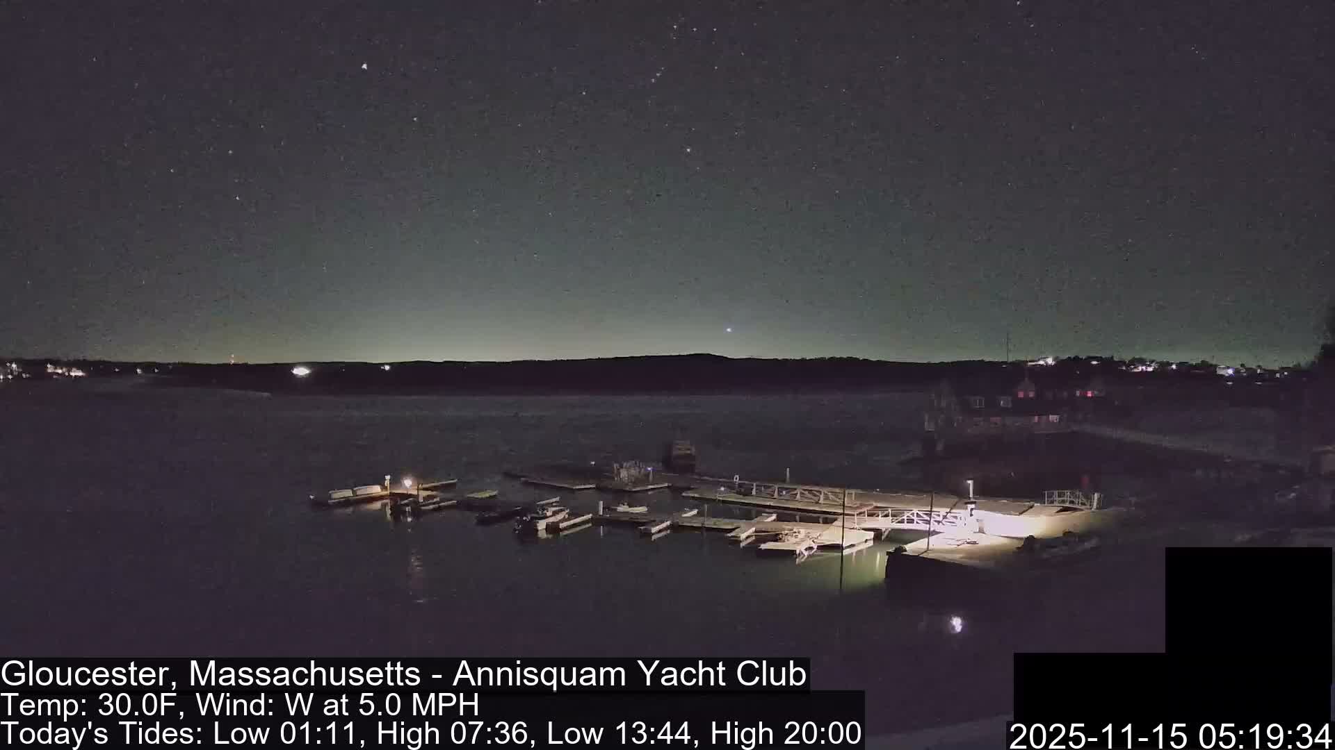 A clear, star-filled night sky hangs over a dark body of water, featuring illuminated docks with several boats and a distant shoreline dotted with lights.