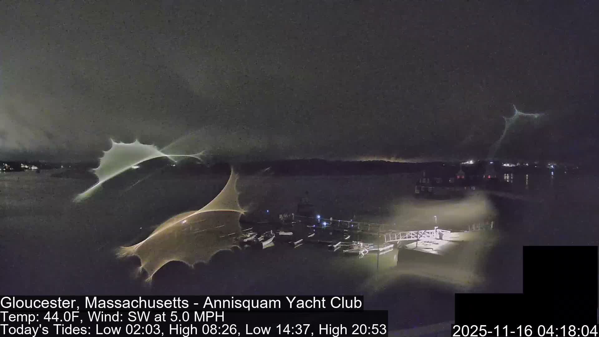 A nighttime view shows several boats moored in a dark harbor with distant shore lights, while prominent bright, irregular light patterns partially obscure the foreground.