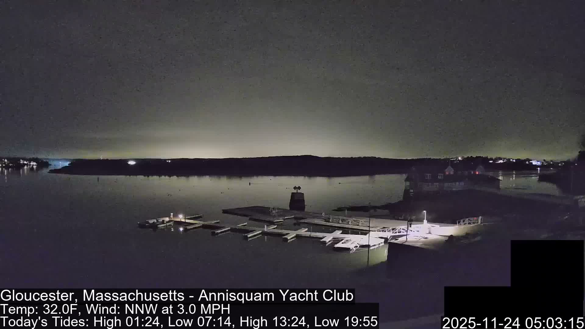 A dark, overcast night settles over a calm body of water featuring illuminated docks with several boats, and distant shorelines twinkling with lights.