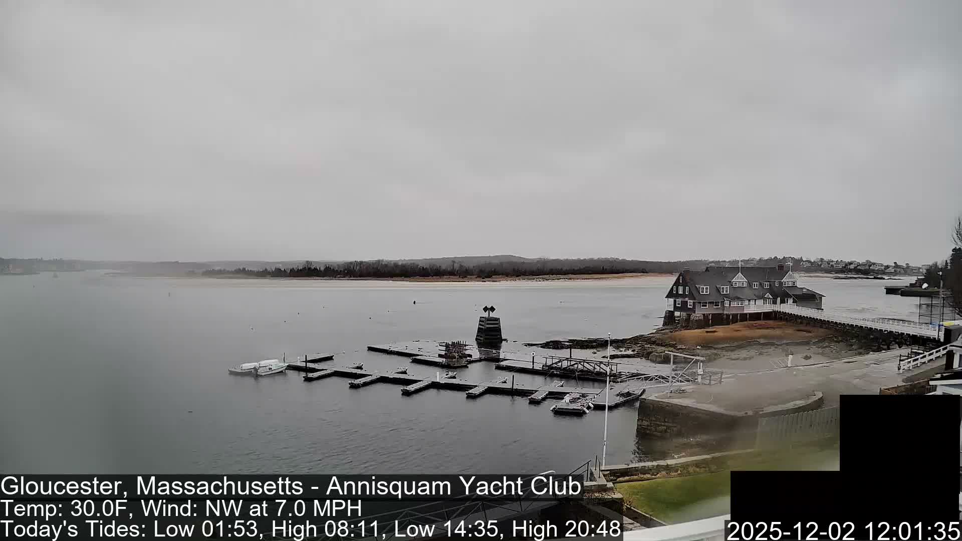 A wide, overcast shot reveals a gray body of water with a large clubhouse building on a rocky shore connected by piers, alongside numerous empty boat docks and a distant, tree-lined landmass.