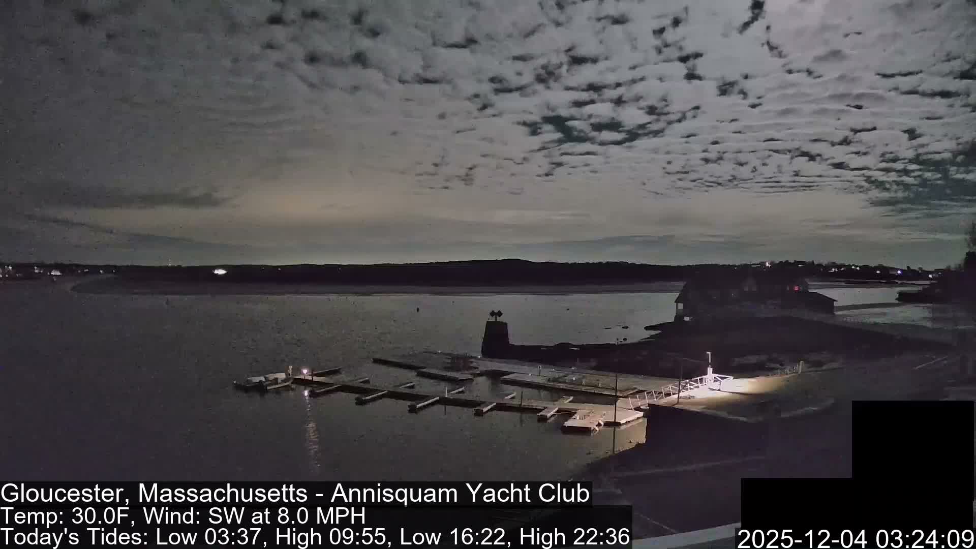 A tranquil nighttime harbor scene displays empty docks and buildings along the shore under a sky filled with scattered clouds.