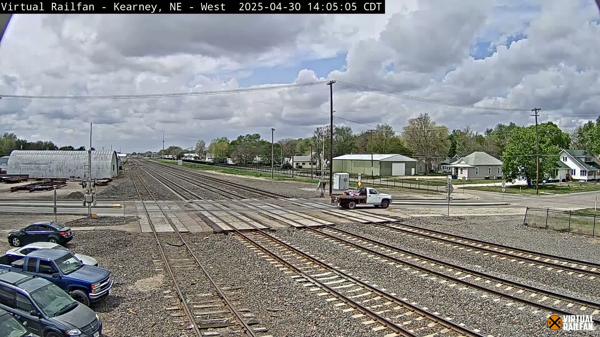 A mostly sunny day shows a railroad crossing in a small town with several vehicles parked near the tracks, and a pickup truck crossing the tracks.