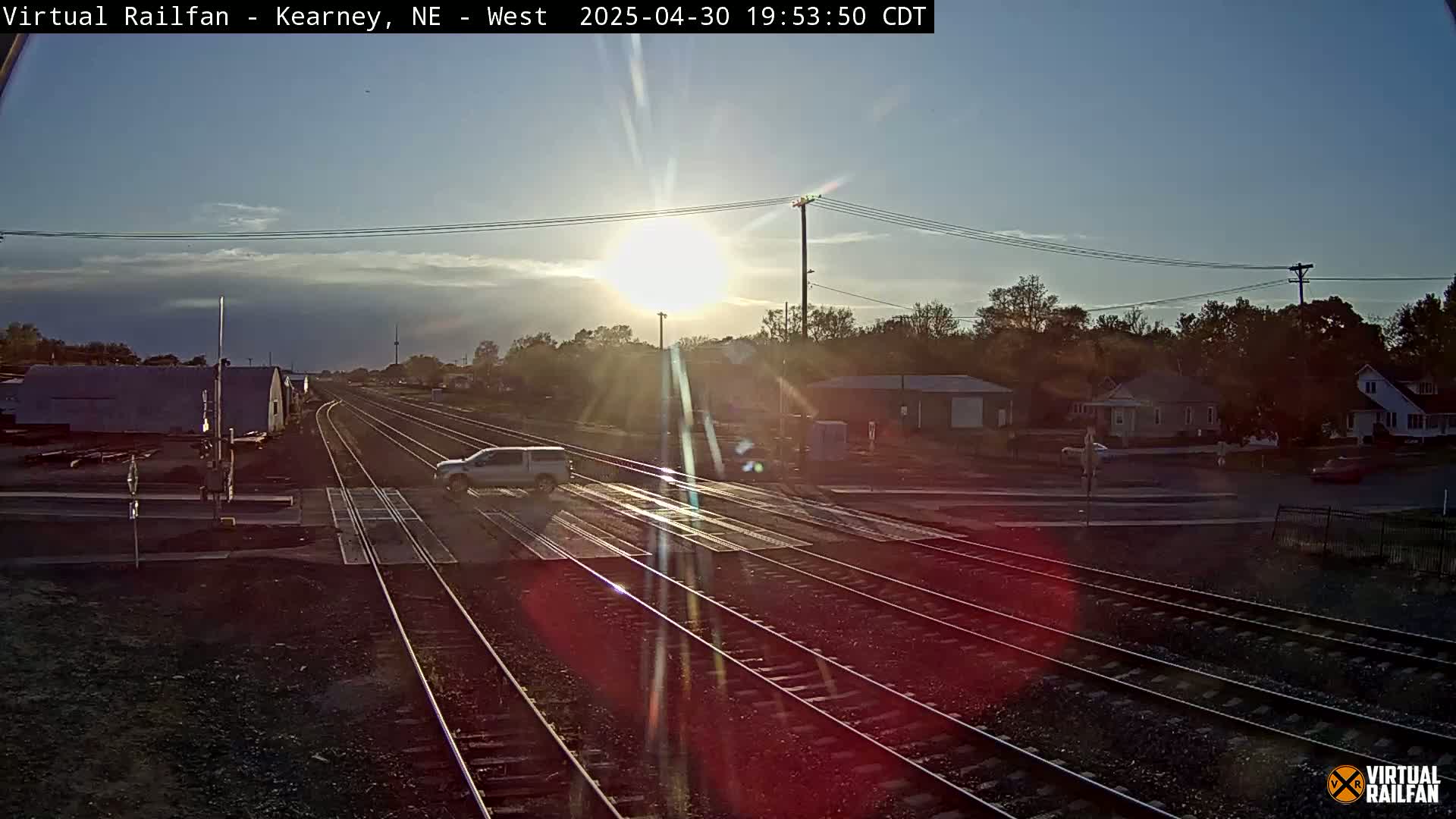 A low sun shines over a railroad crossing in a residential area, with a light-colored vehicle crossing the tracks.