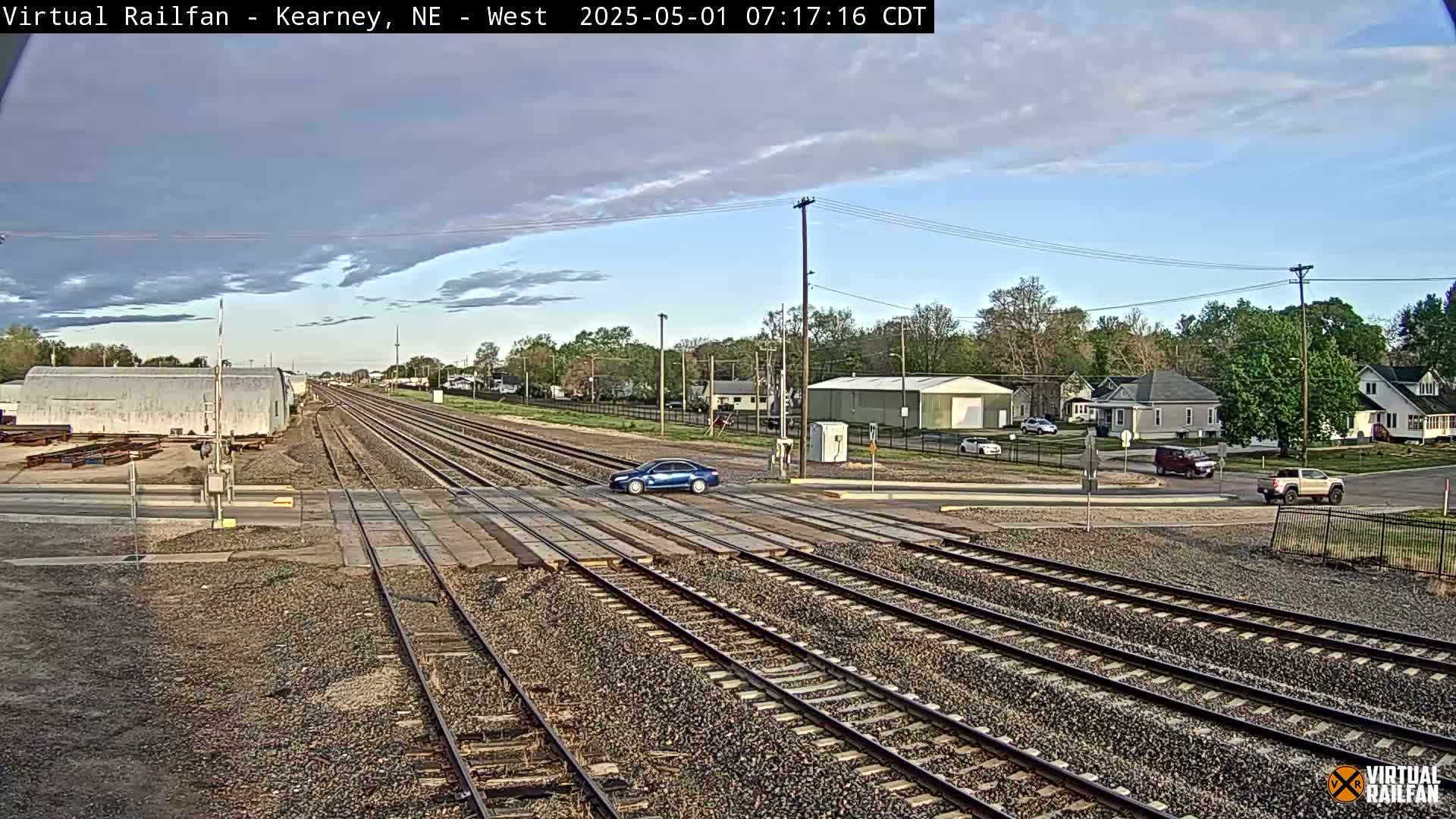 Multiple train tracks intersect a road with several cars at a crossing, under a partly cloudy sky.