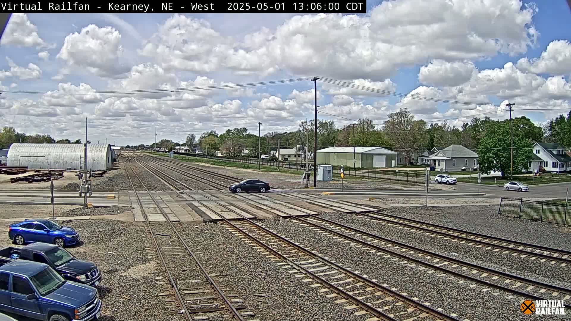 A partly cloudy sky hangs over railroad tracks crossing a street in a residential area, with several cars visible at the crossing and parked nearby.