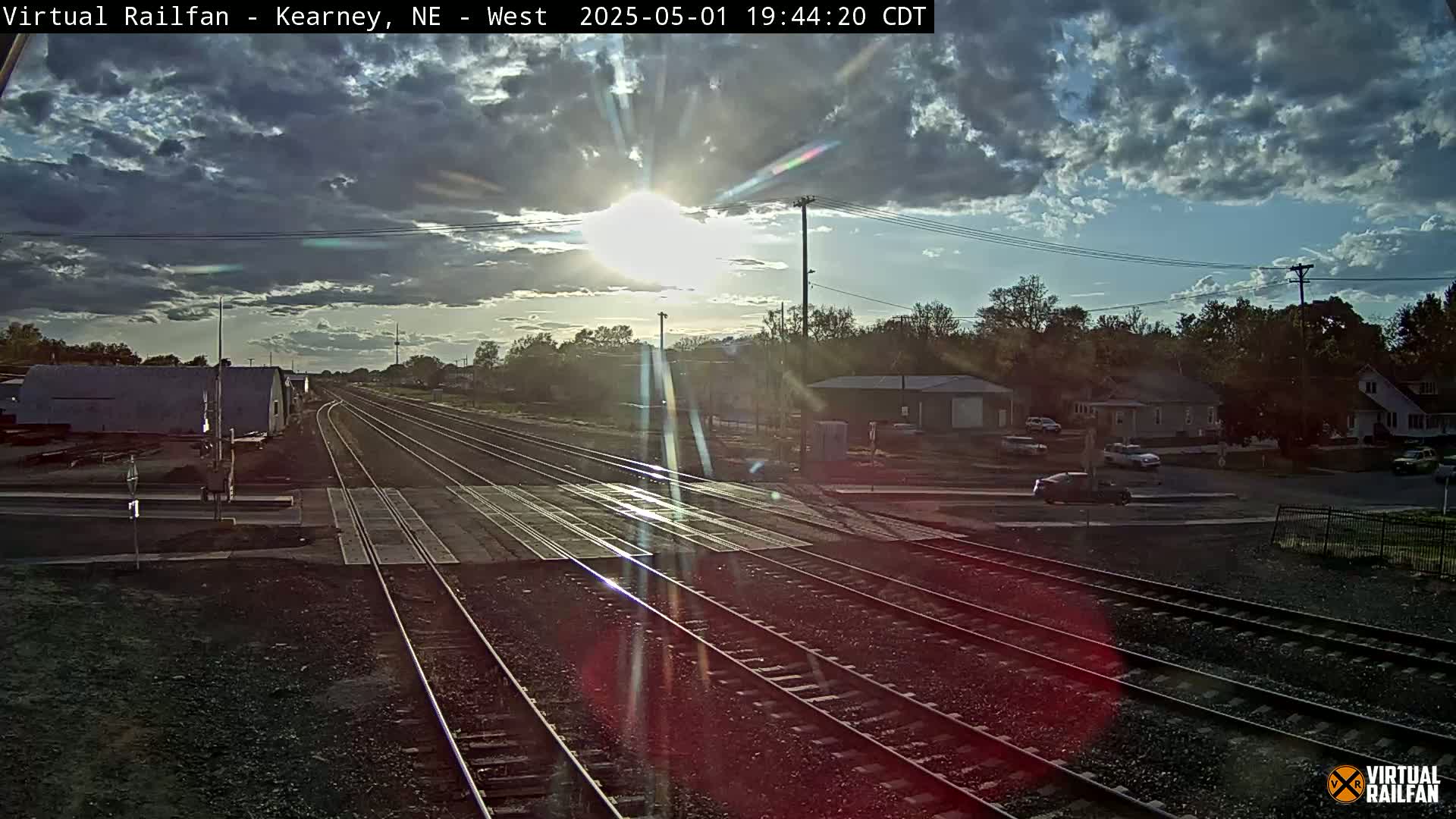 A mostly cloudy sunset illuminates a railroad crossing with several tracks, flanked by buildings and cars.