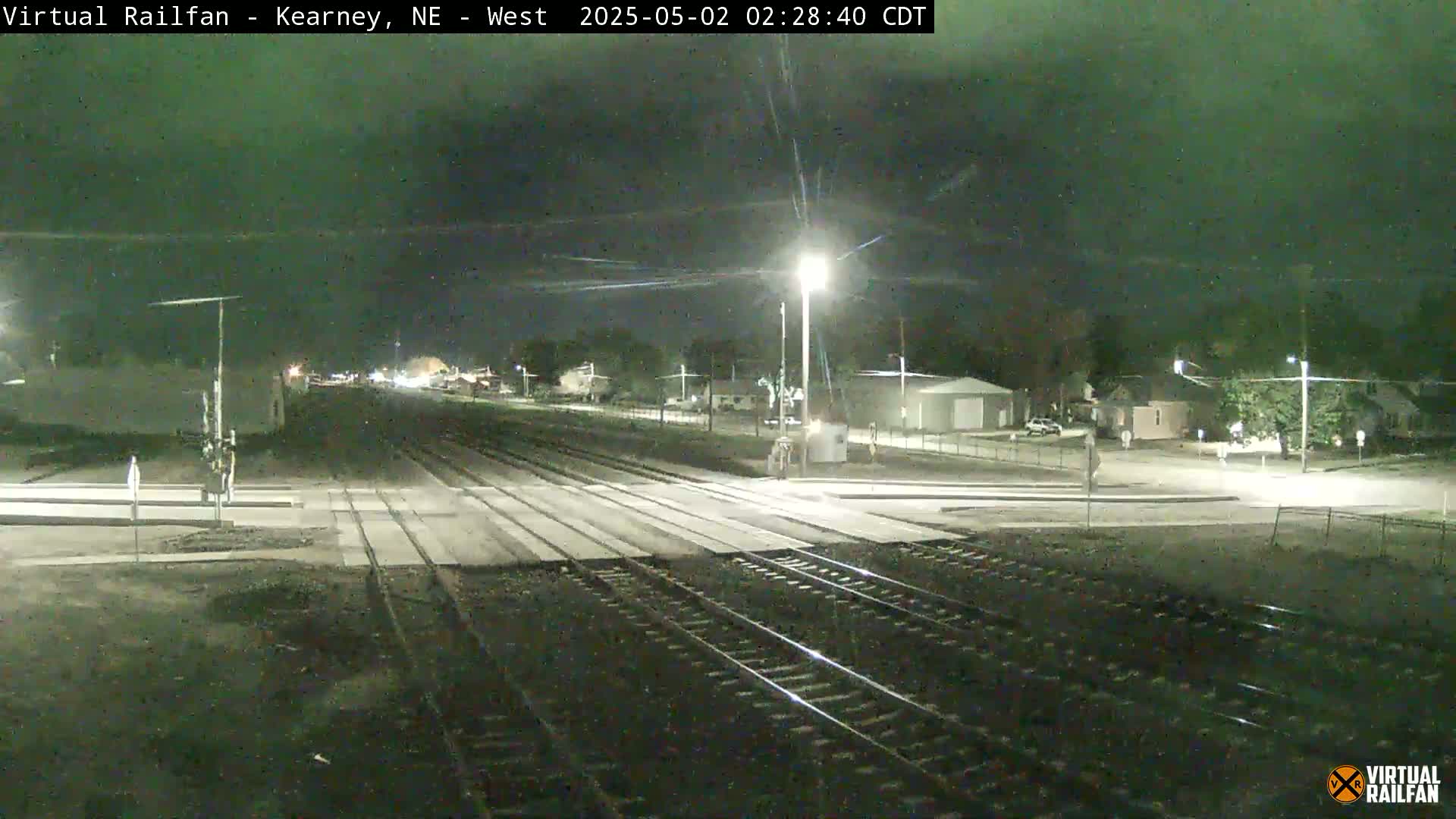 A nighttime view of railroad tracks intersecting a road, with a few buildings and streetlights visible in the background under a dark sky.