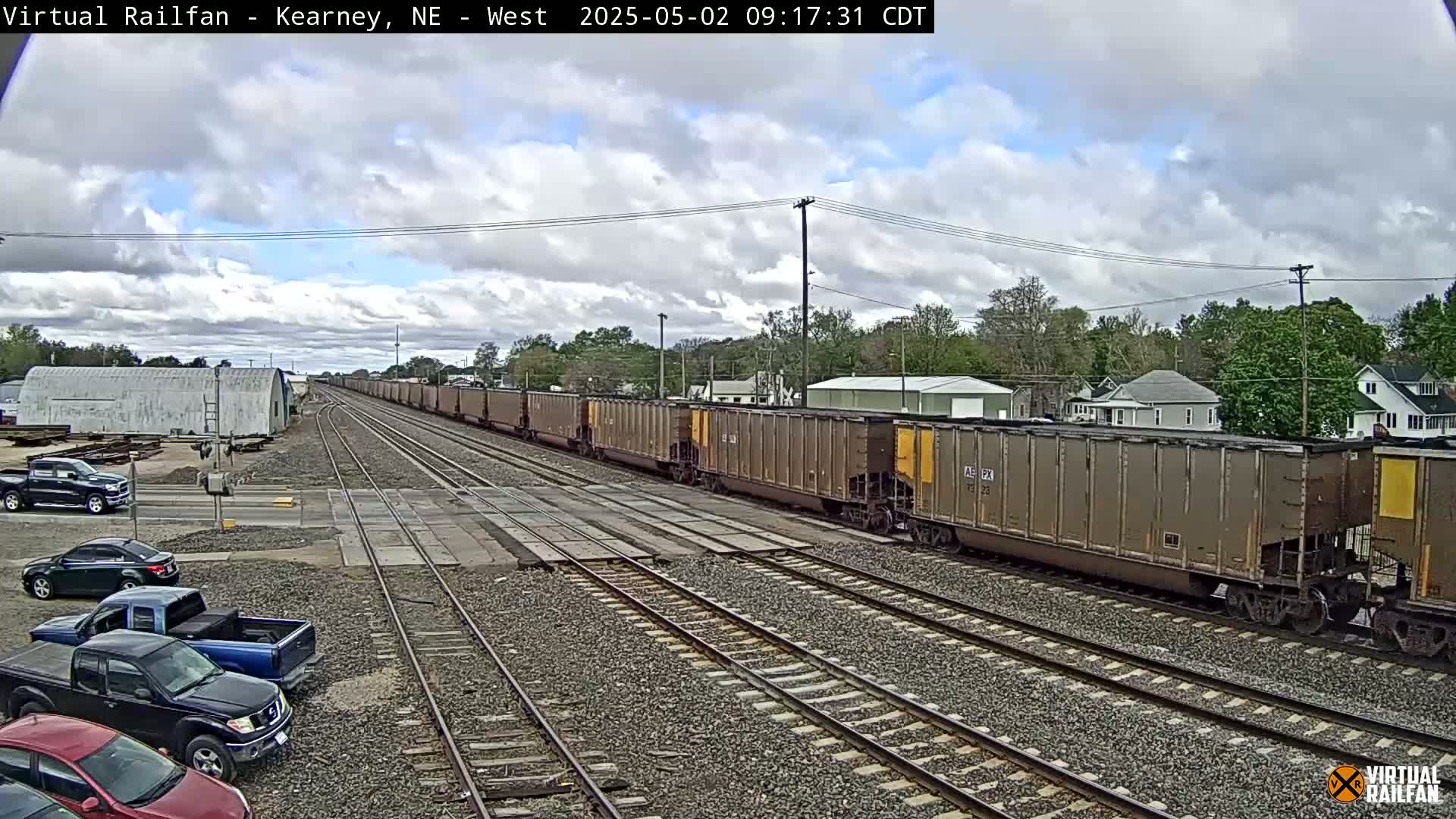 A long freight train passes through a railway crossing under a partly cloudy sky, with several parked pickup trucks visible in the foreground.
