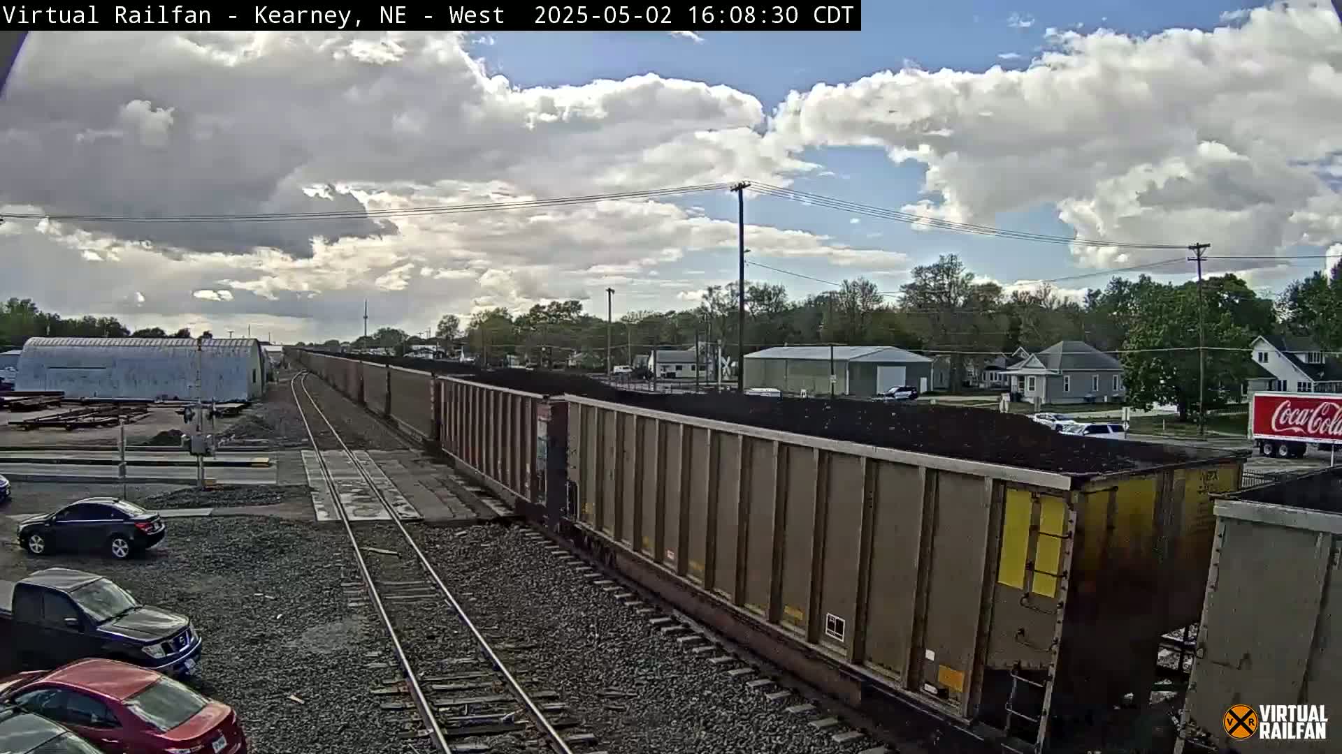 A long train carrying coal sits on railroad tracks under a partly cloudy sky, near a road with cars and houses in the background.