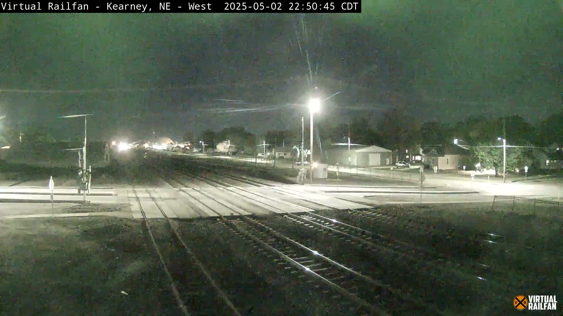 A nighttime view of a railroad crossing in a residential area under a clear sky, showing train tracks intersecting a road with streetlights illuminating the scene.