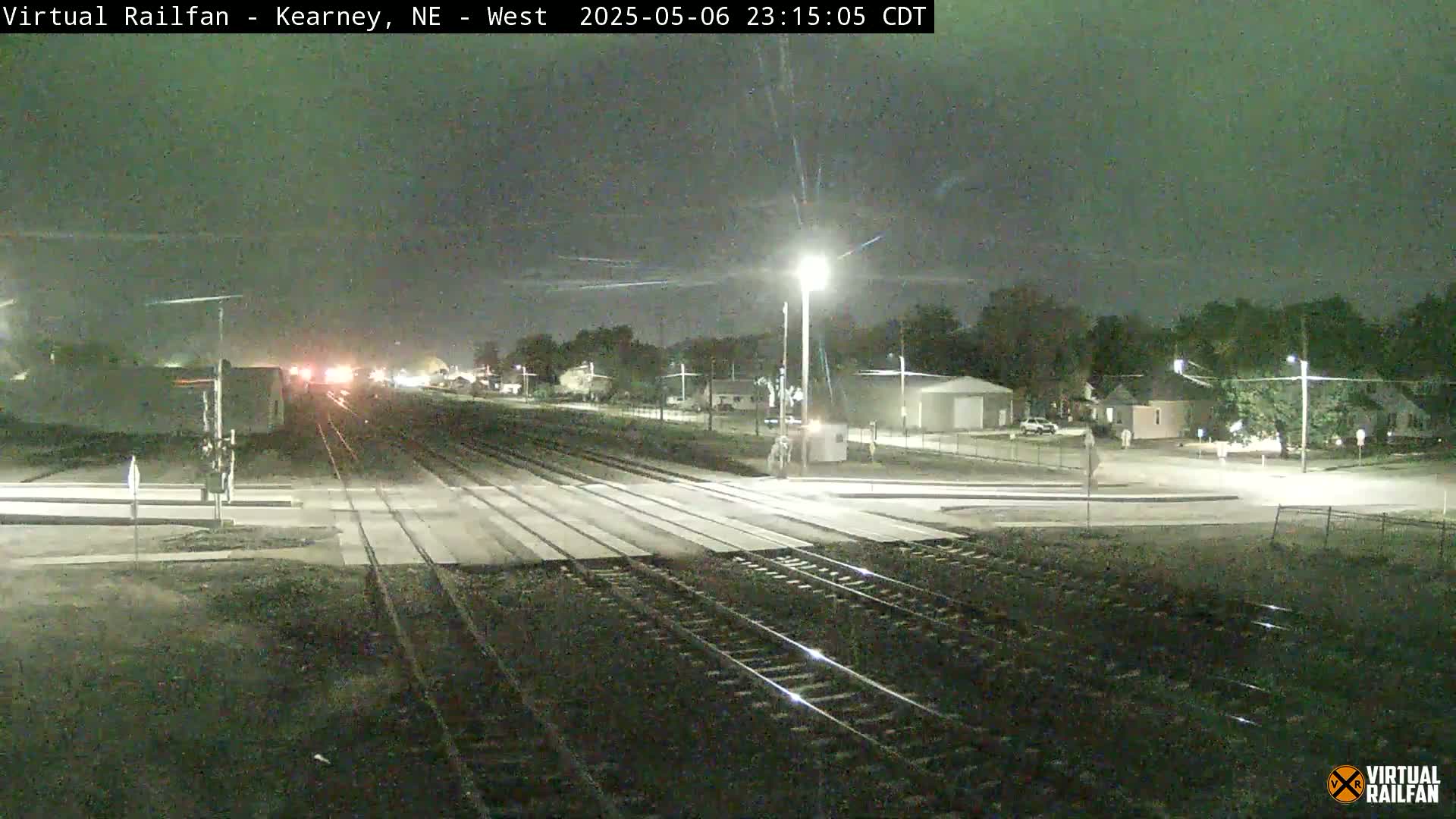 A nighttime view of railroad tracks intersecting a road, with dimly lit buildings and trees in the background under a dark sky.