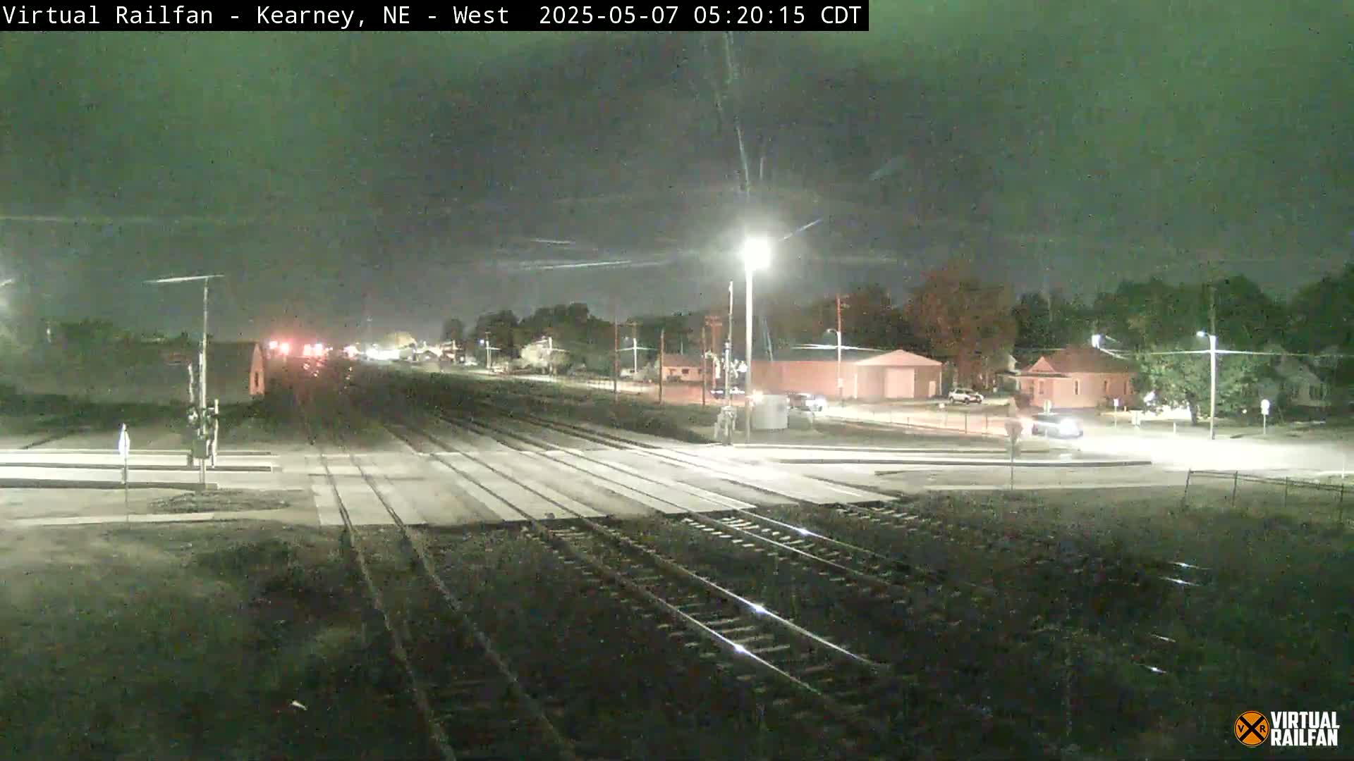 A nighttime view of a railroad crossing with a few vehicles at a nearby intersection under a partly cloudy sky.
