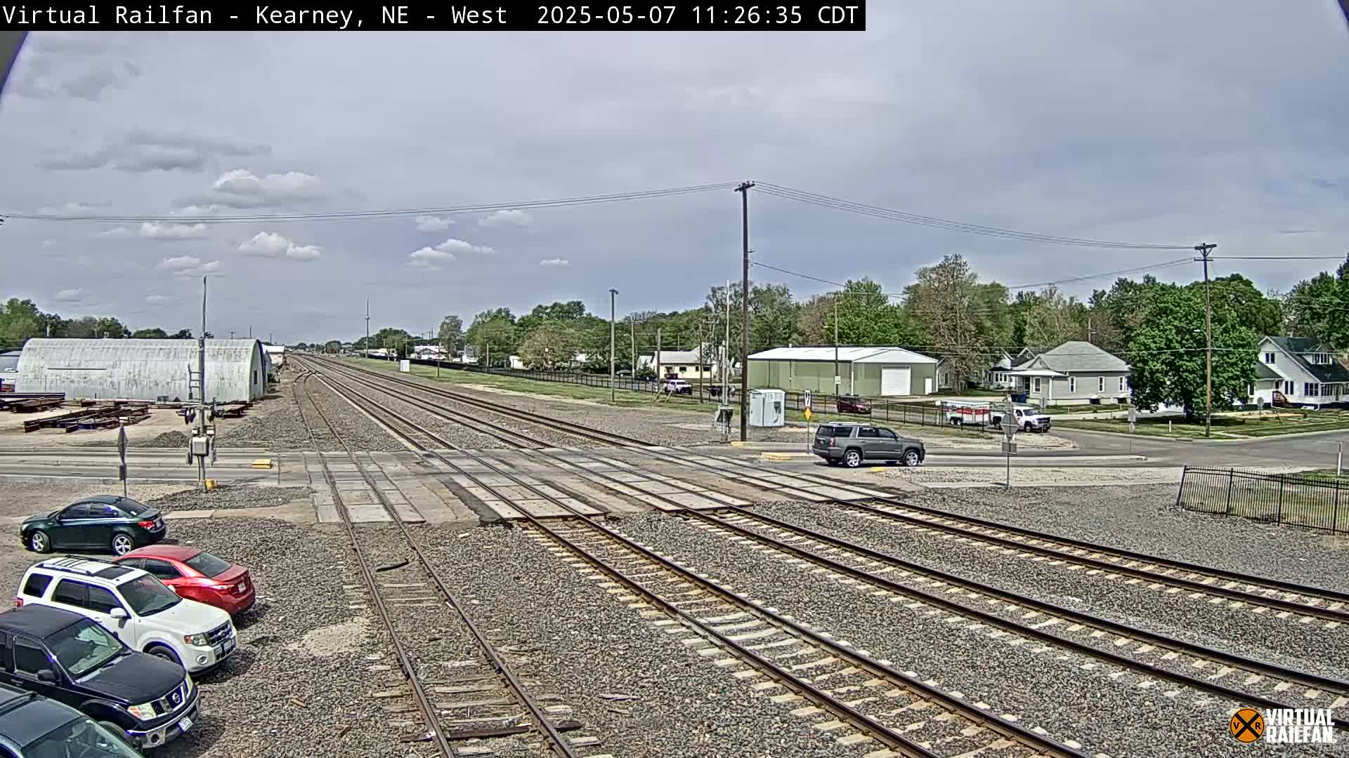 Under a partly cloudy sky, railroad tracks cross a road in front of a residential area with several parked cars visible near the tracks.