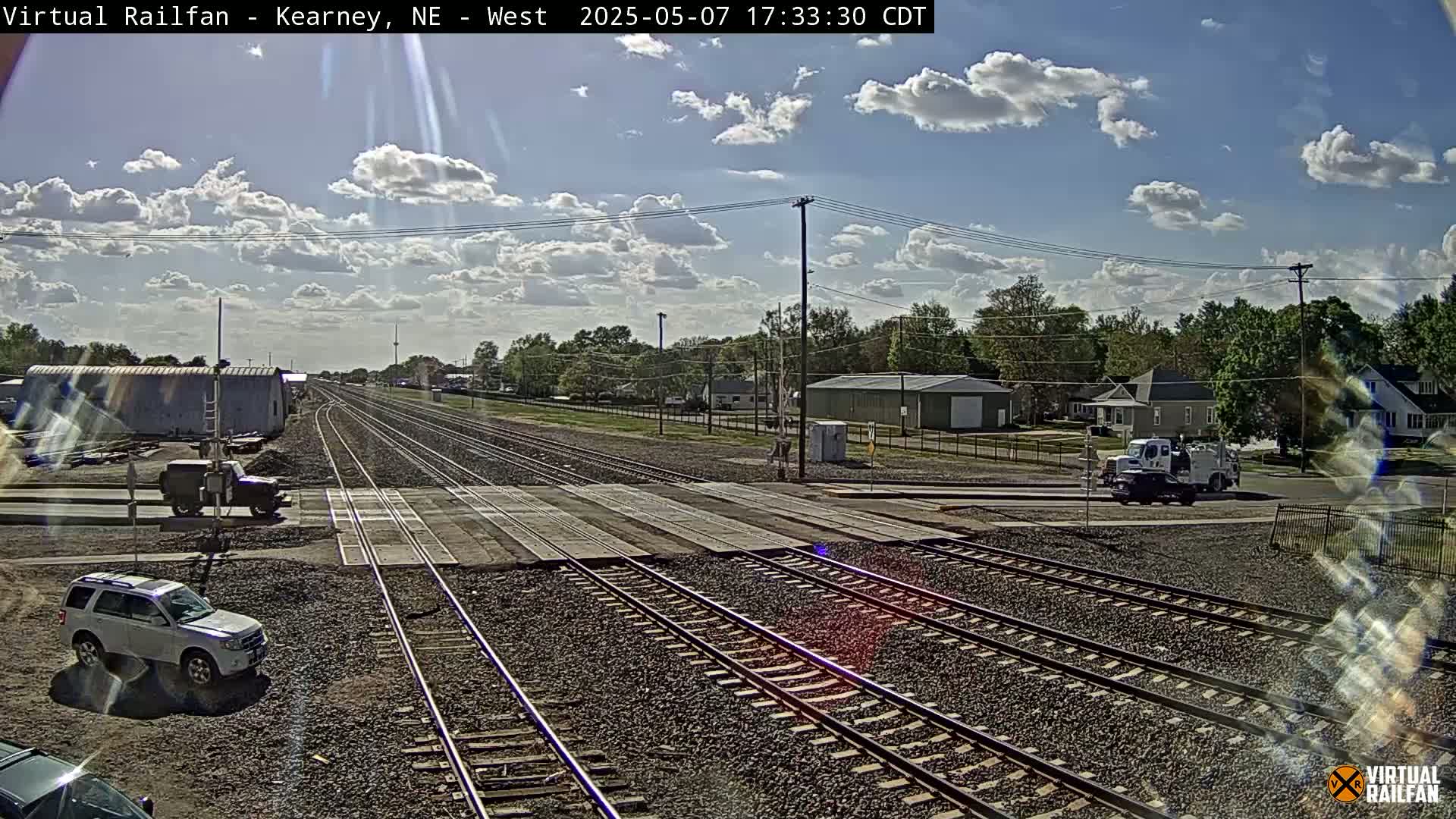 A sunny day with partly cloudy skies shows a railroad crossing with several sets of train tracks, a few vehicles, and buildings in the background.