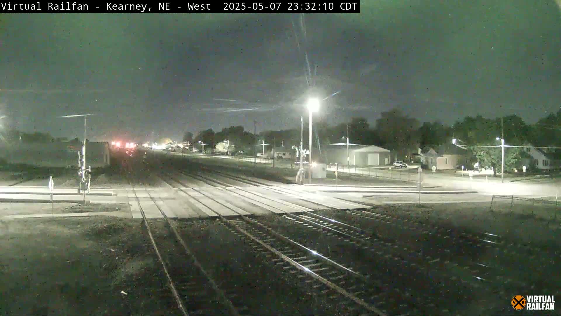 A nighttime view of a railroad crossing with a street intersecting, illuminated by streetlights under a clear, dark sky.