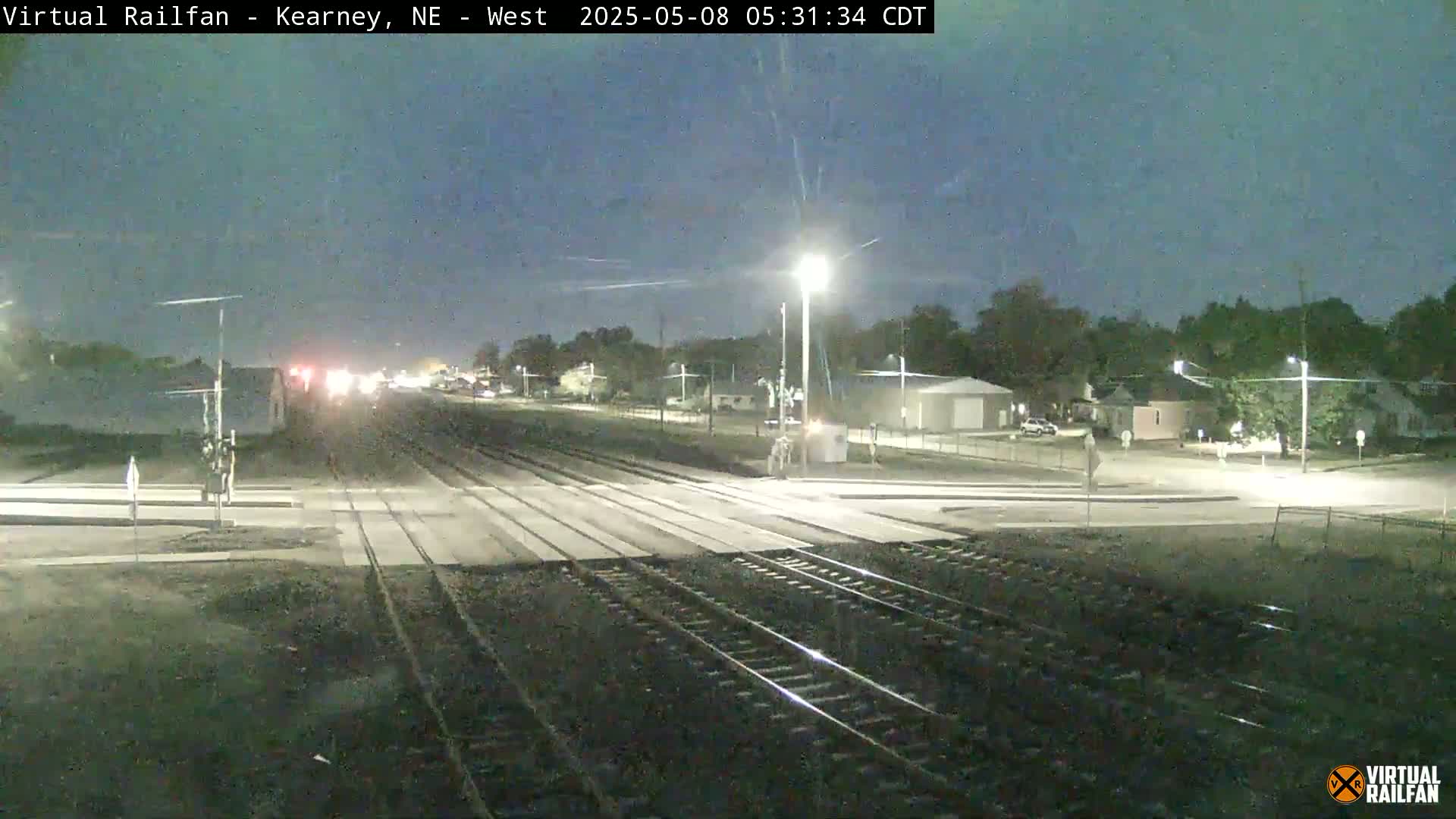 A nighttime view of a railroad crossing in a residential area, under a dark sky.