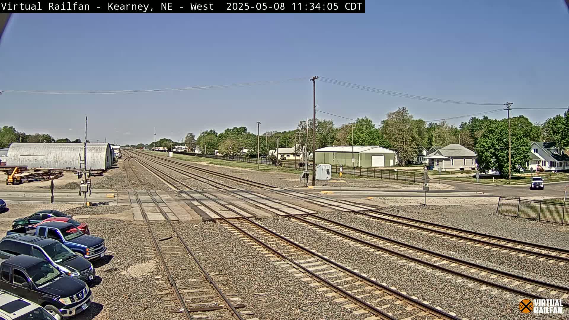 A railroad crossing in a residential area on a sunny day, with several cars parked nearby and a few houses and buildings visible in the background.