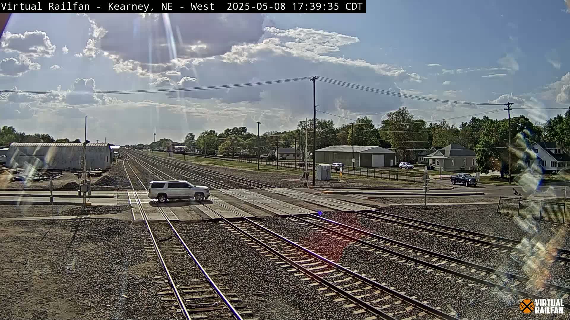 A partly cloudy sky illuminates a mostly sunny scene of a railroad crossing where a silver SUV is stopped at the tracks, with residential buildings and trees in the background.